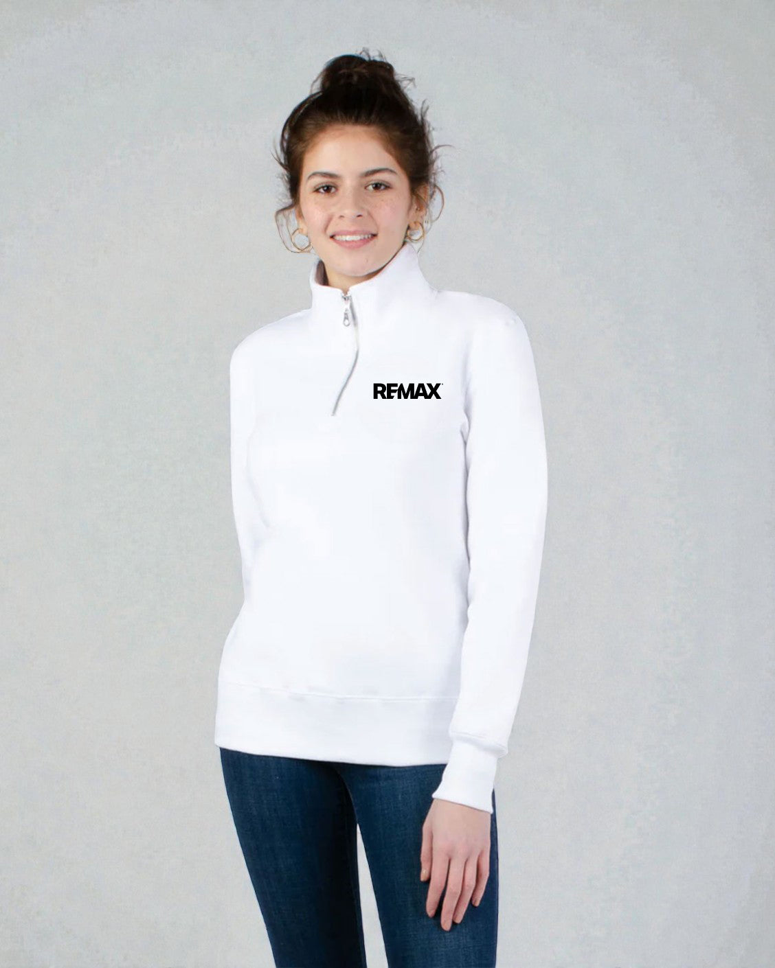 A woman wearing a white long-sleeve quarter-zip pullover sweatshirt with the black REMAX logo on the left chest, paired with dark blue jeans, standing against a textured light gray background.