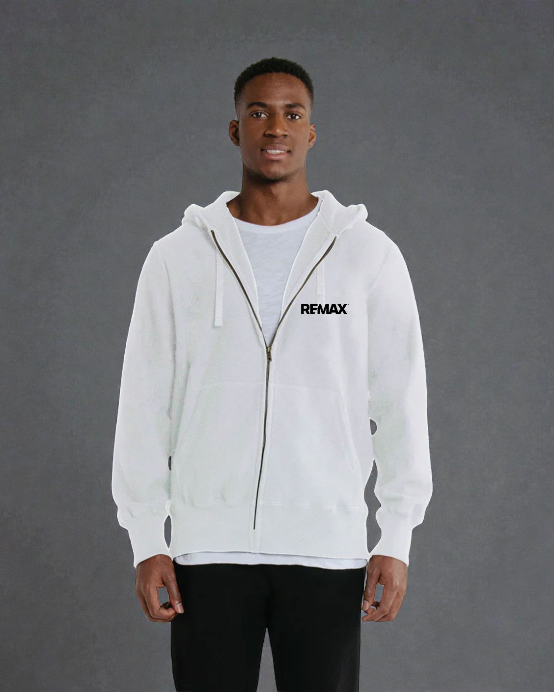 A man wearing a white zip-up hoodie with a black REMAX logo on the left chest, layered over a light gray t-shirt and black pants, standing against a textured gray studio background.