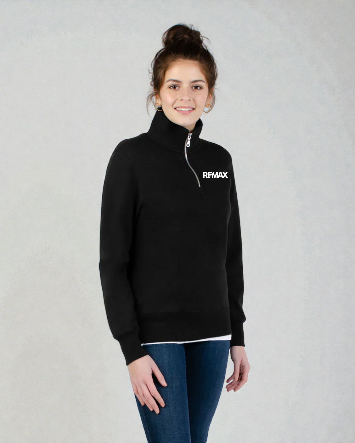 A woman wearing a black quarter-zip mock neck sweater with a white 'RFMAX' logo on the chest, paired with blue jeans, standing against a light gray textured background.