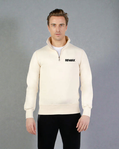 A man wearing a cream-colored quarter-zip sweatshirt with a black REMAX logo on the left chest, paired with black pants, standing against a neutral gray studio background.