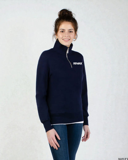 A woman wearing a navy blue quarter-zip pullover sweater with a white 'RFMAX' logo on the left chest, paired with dark blue jeans, standing against a light gray textured background.
