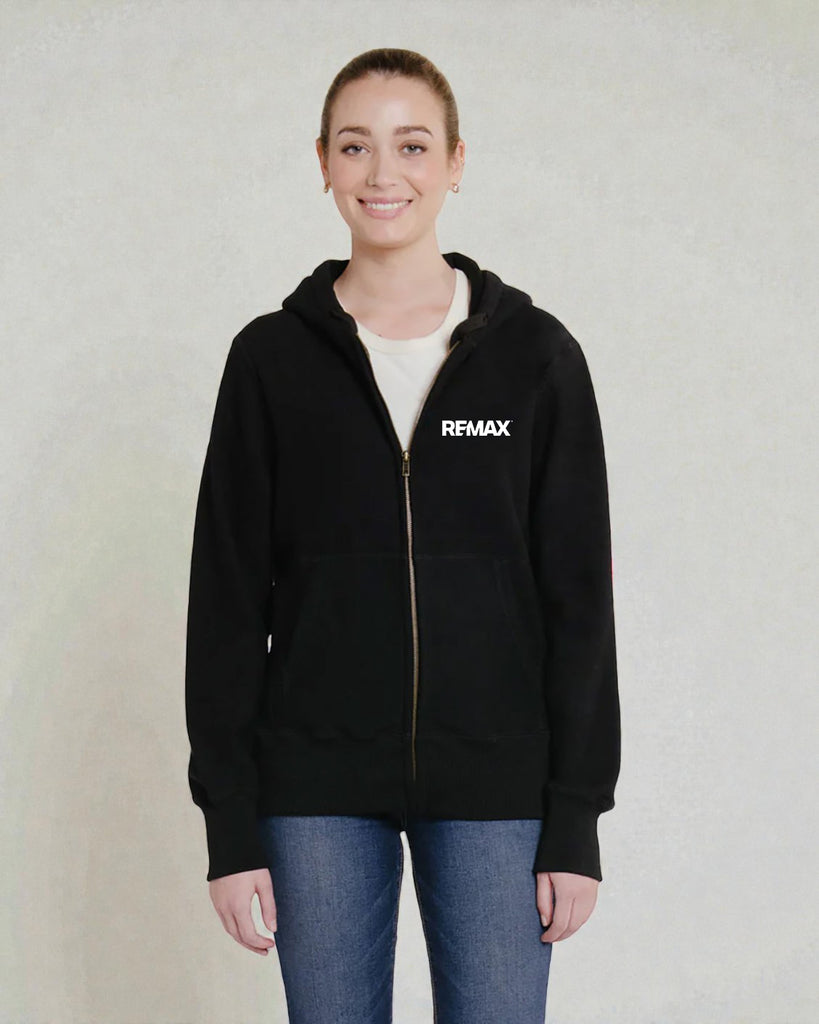 A woman wearing a black zip-up hoodie with a white REMAX logo on the left chest, paired with a white t-shirt and blue jeans, standing against a textured light gray wall.