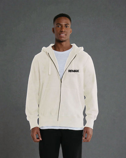 A man wearing a cream-colored zip-up hoodie with a black REMAX logo on the left chest, layered over a light blue t-shirt and black pants, standing against a dark gray studio background.