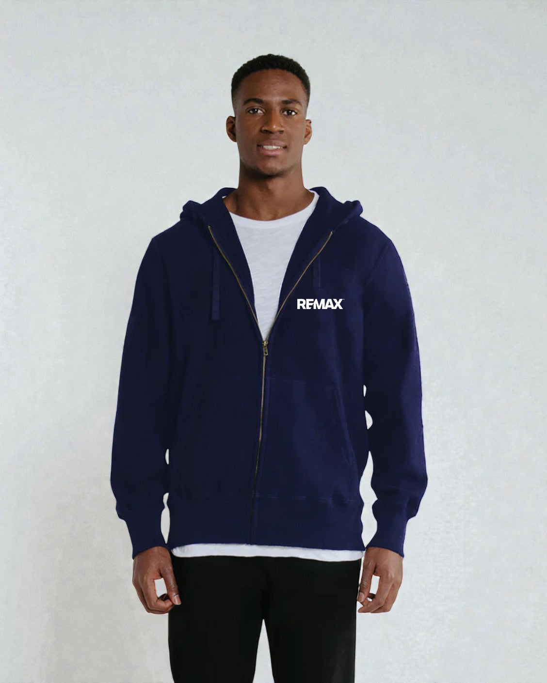 A man wearing a navy blue zip-up hoodie with a white REMAX logo on the left chest, layered over a white t-shirt and black pants, standing against a light gray textured wall.