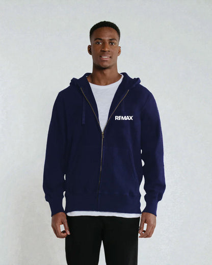 A man wearing a navy blue zip-up hoodie with a white REMAX logo on the left chest, layered over a white t-shirt and black pants, standing against a light gray textured wall.