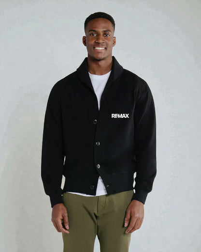 A man wearing a black button-up varsity-style jacket with a white 'RFMAX' logo on the chest, layered over a white t-shirt and paired with olive green pants, standing against a light gray textured wall.