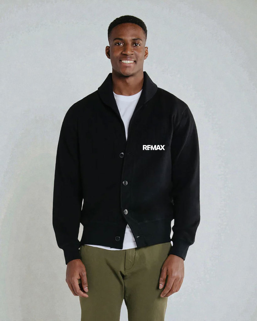 A man wearing a black button-up varsity-style jacket with a white 'RFMAX' logo on the chest, layered over a white t-shirt and paired with olive green pants, standing against a light gray textured wall.