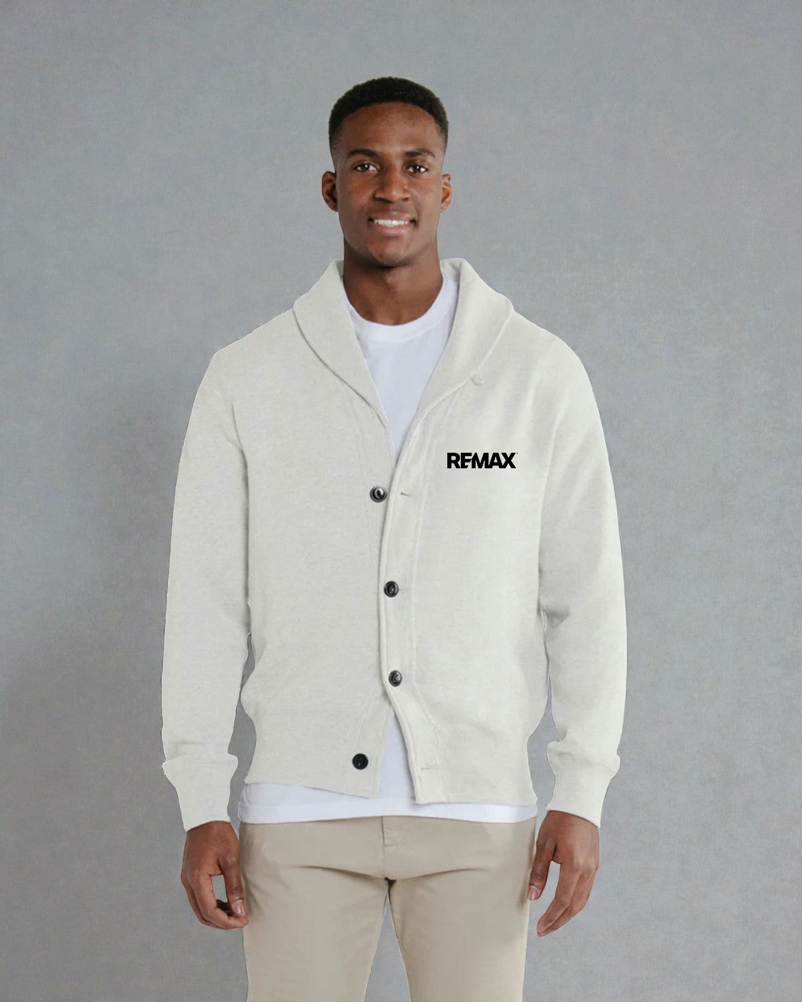 A man wearing a light cream-colored cardigan with a shawl collar, black buttons, and a black 'RFMAX' logo on the left chest, styled over a white t-shirt and beige pants against a gray studio background.