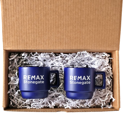 Two blue mugs with 'REMAX Stonegate' text in a cardboard box with shredded paper.