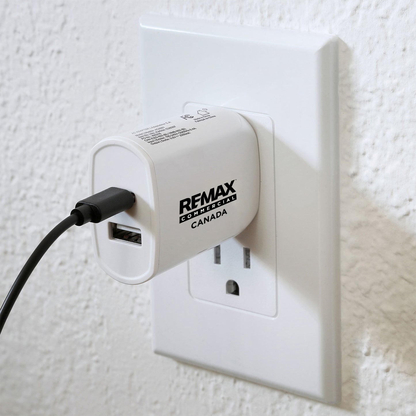 A white REMAX PowerWall travel adapter plugged into a white wall outlet, with a black power cord extending to the left against a textured white wall.